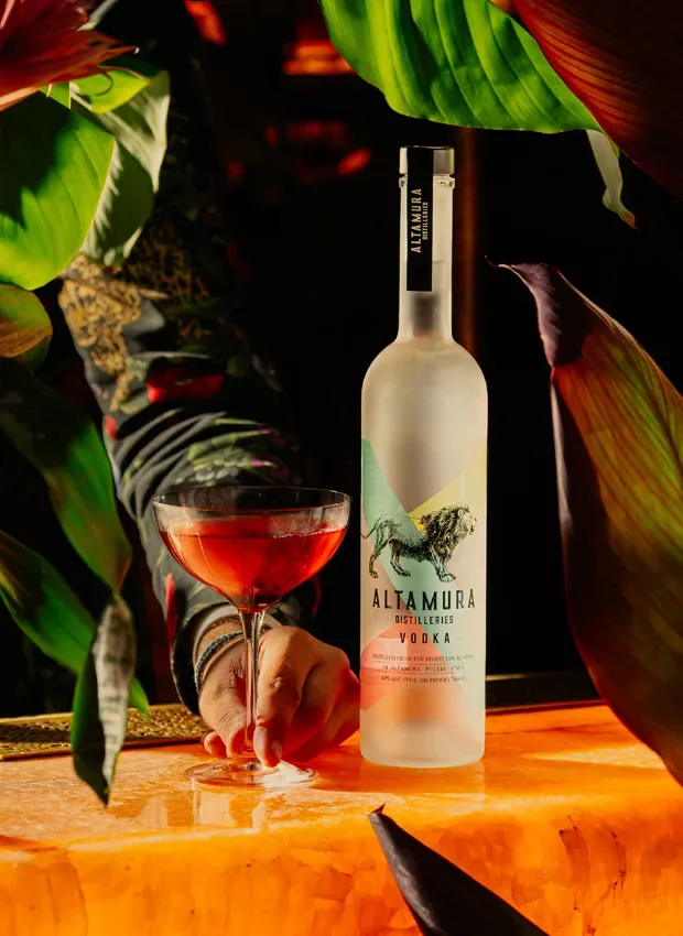 Garrafa turquesa de Altamura Distilleries Vodka ao lado de um coquetel rosa, entre folhagem tropical