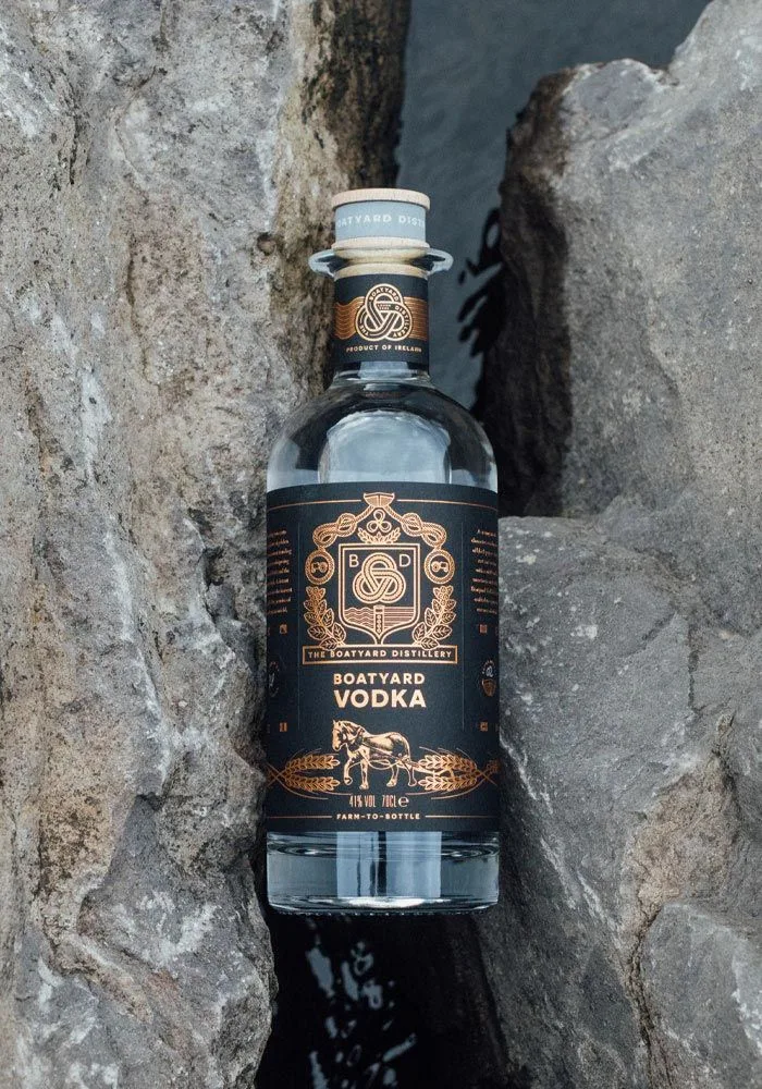 Garrafa preta e cobre de Boatyard Vodka encaixada entre rochas, com o rótulo do cavalo em destaque